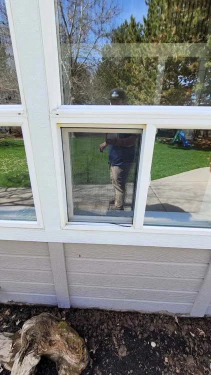 A person reflected in a window, standing outside with a yard and trees visible.