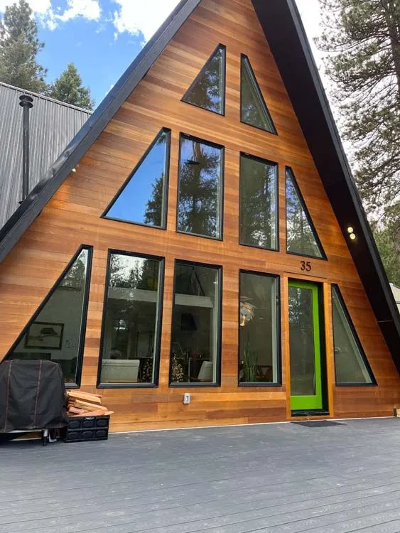 A-frame house with brown wooden siding, black trim, and a green door. Tall windows reflect trees.