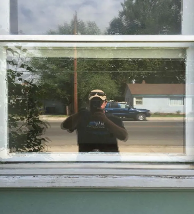 Reflection in window shows person holding phone, wearing a hat and shirt, with a street and houses in background.