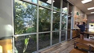 Large windows overlooking a green outdoor area. A person cleans inside near tables and an exit.